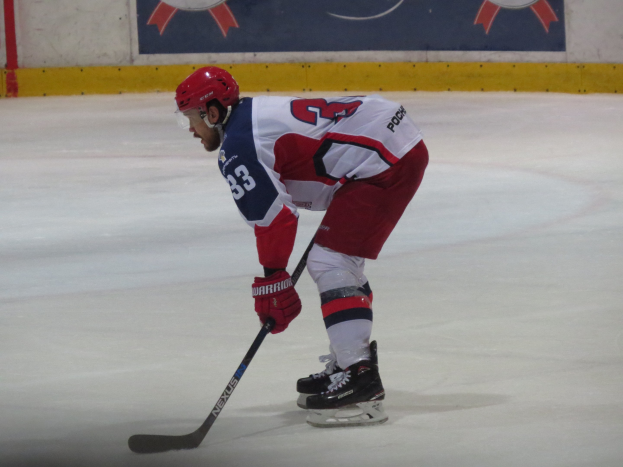 Eishockey-Spieler in voller Ausrüstung auf dem Eis mit einem Banner im Hintergrund, das Text und Logos zeigt.