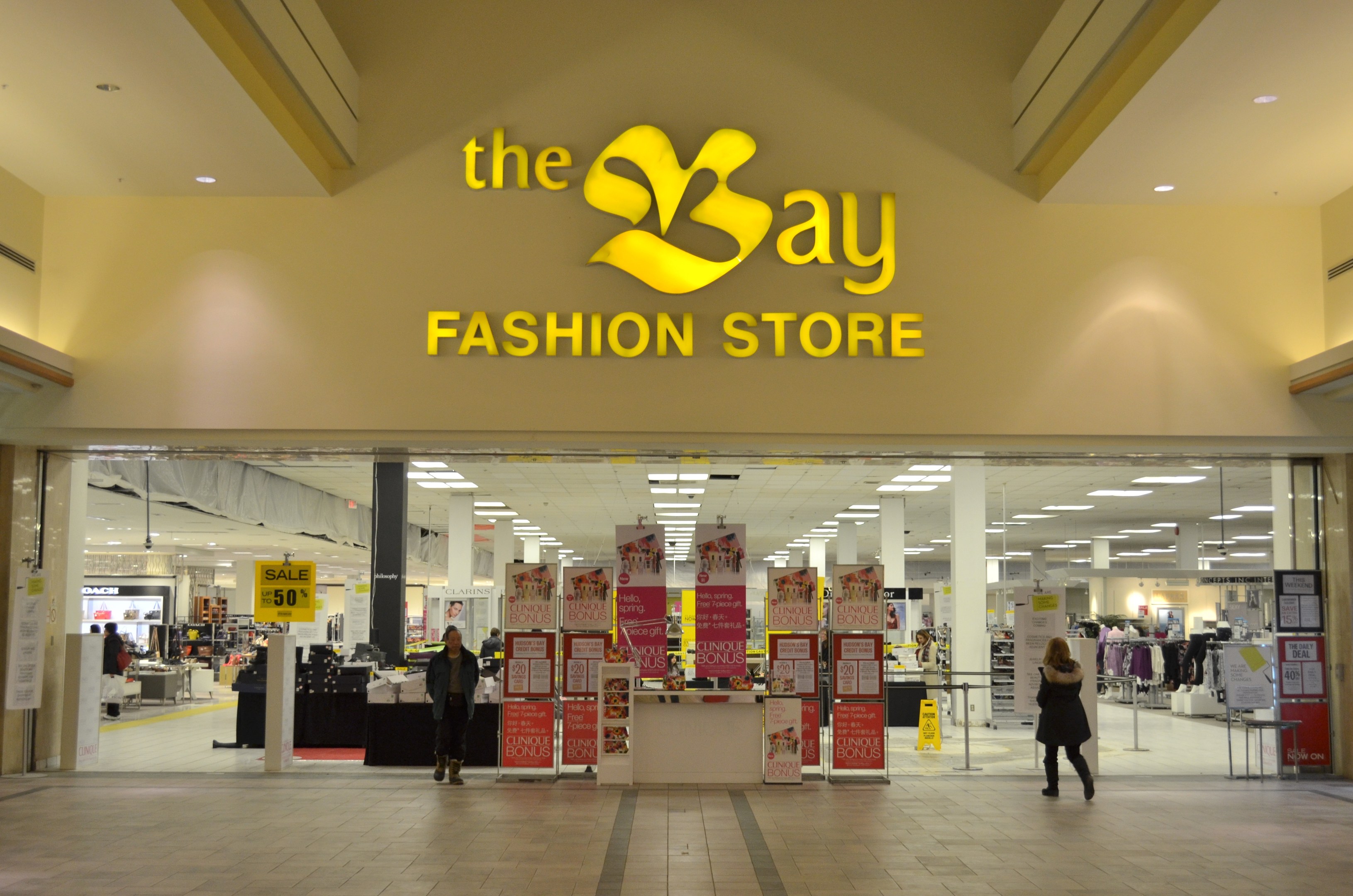 Ein Modegeschäft namens "bay fashion" in einem Einkaufszentrum mit Einkäufern, Wandtextanzeigen, Säulen, Deckenbeleuchtung und Hintergrunddekoration.