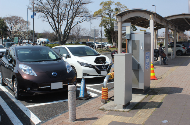 Elektroauto-Ladestation in Japan mit Autos, Verkehrskegeln, einer Person auf dem Gehweg, einem Schuppen, Masten, Lichtern, Schildern, Bäumen, Pflanzen und einem Himmel im Hintergrund.