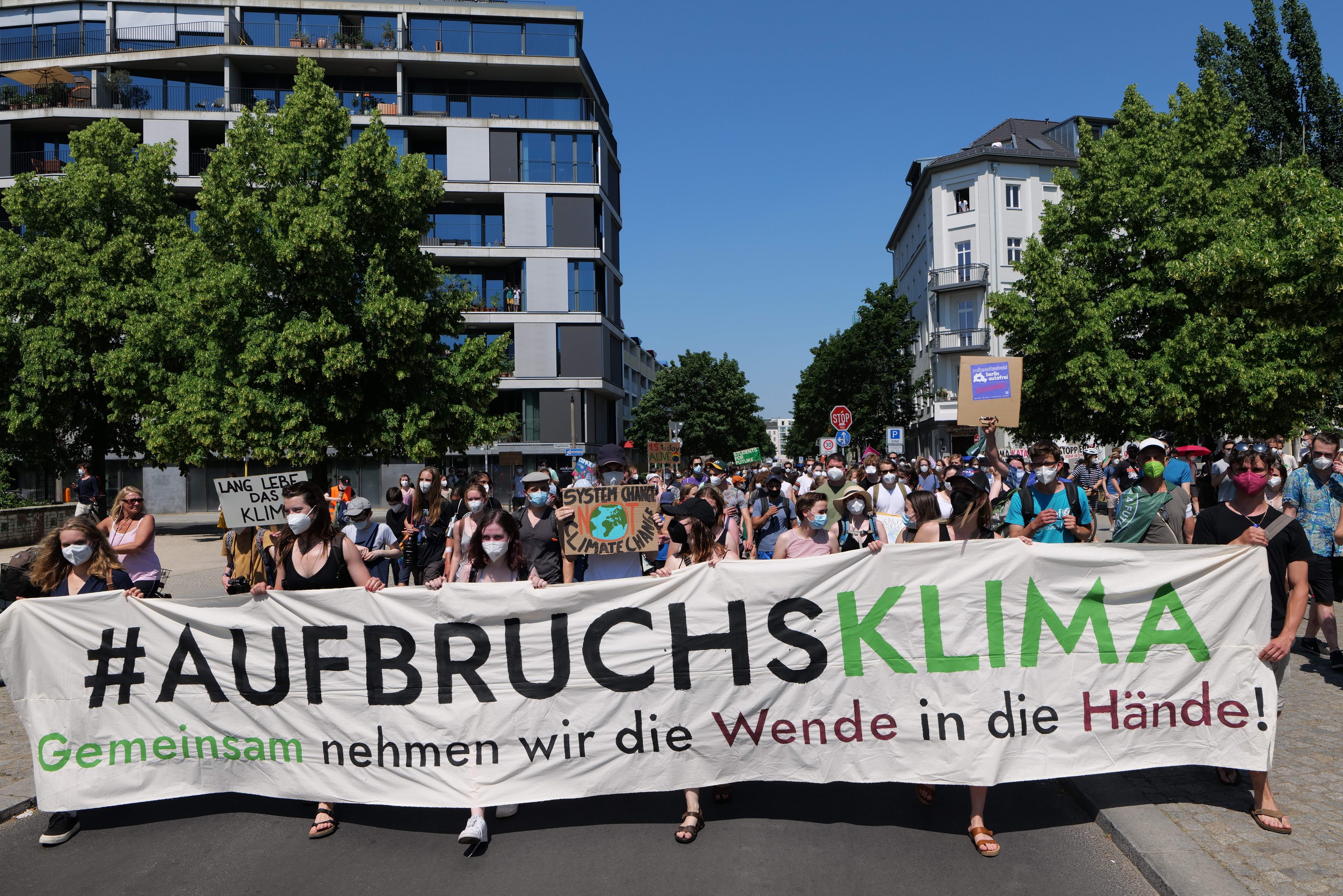 Gruppe von Menschen mit Masken, die ein Banner mit der Aufschrift "Aufbruchsklima" vor einem Gebäude mit Bäumen und wolkenlosem Himmel halten.