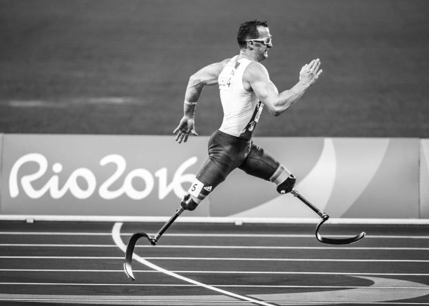 Schwarzes und weißes Bild eines Mannes mit einem Prothesenbein, der auf einer Laufbahn läuft, mit einer Tafel im Hintergrund, auf der "Rio 2016 Paralympische Spiele" steht.