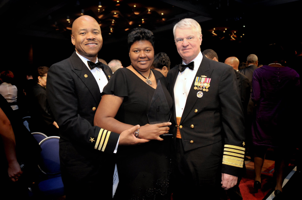 Zwei Männer in schwarzen Blazern und weißen Hemden stehen neben einer Frau in einem schwarzen Kleid, die eine Trophäe auf einem roten Teppich hält, während andere im Hintergrund sitzen und stehen und von beleuchteten Decken umgeben sind.