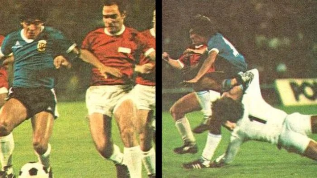 Zwei Männer in Fußballtrikots kämpfen um den Ball während eines Spiels auf einem Feld mit einer Stadiontafel im Hintergrund.