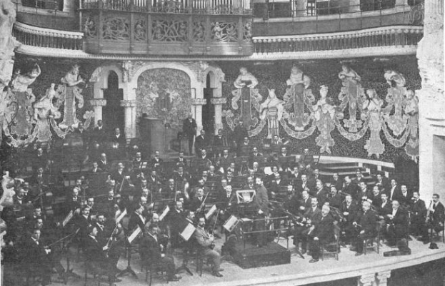 Ein Schwarz-Weiß-Foto eines großen Orchesters in einem Auditorium, mit Musikern, die auf der Bühne sitzen und stehen, einem Podium in der Mitte und Skulpturen an den Wänden.