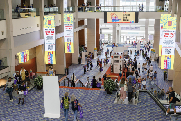 Große Gruppe von Menschen, die durch ein großes Gebäude mit Bannern, Topfpflanzen, Rolltreppen, Geländern, Säulen, bedruckten Schildern, Glastüren und anderen Gegenständen geht, wobei einige Taschen tragen, auf der Nationalen Buchmesse.