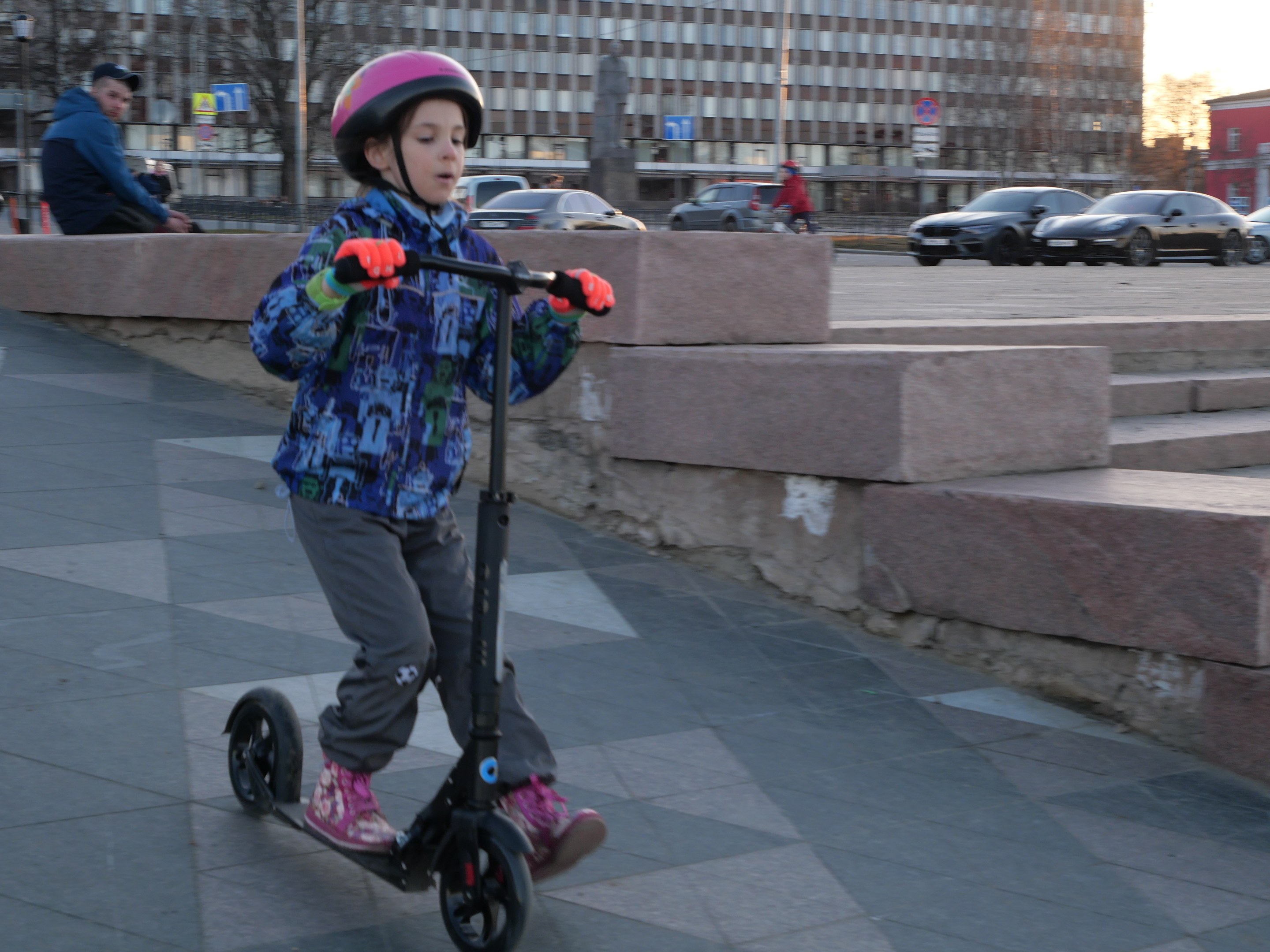 Ein junger Junge mit Helm und Handschuhen fährt auf einem Roller eine Treppe auf dem Gehweg hinunter, mit Fahrzeugen, Menschen, Bäumen, Polen, Brettern, Gebäuden und einem klaren blauen Himmel im Hintergrund.