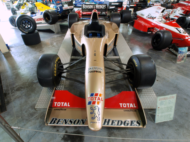 Eine Gruppe von Rennwagen, die hinter einem Metallzaun in einem Motorsportmuseum ausgestellt sind, mit einer Texttafel und Menschen im Hintergrund.