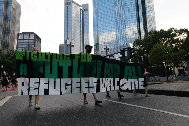 Eine Gruppe von Menschen marschiert mit einem Banner, auf dem "Kämpfen für eine Zukunft für alle - Willkommen Flüchtlinge" steht, die Straße entlang vor Bäumen, Gebäuden und Ampeln unter einem bewölkten Himmel.