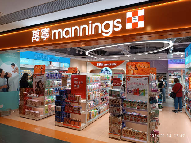 Ein Manning's-Laden in einem Einkaufszentrum mit Menschen auf dem Boden, einige halten Taschen, Regale mit Gegenständen, Schilder, Deckenbeleuchtung und ein Wasserzeichen.