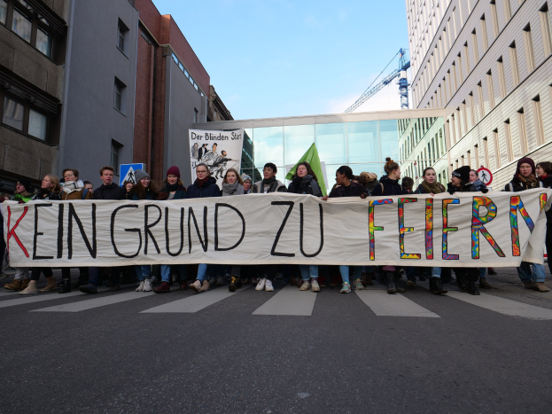 Demonstranten halten ein Banner mit der Aufschrift "Kein Grund zu Feiern" gegen deutsche Sparmaßnahmen, mit Gebäuden, einem Kran und einem bewölkten Himmel im Hintergrund.