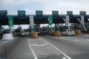 Eine Mautstation an der Seite einer Straße mit darunter fahrenden Autos, umgeben von Verkehrskegeln, Absperrpfählen, Schildern, einem Tor, Laternenmasten, Bäumen, Gebäuden und einem bewölkten Himmel im Hintergrund.