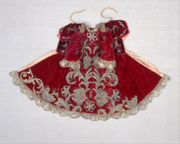 Ein rotes Samtkleid mit goldener Stickerei, das auf einem Bügel an einer weißen Wand hängt und mit detaillierten Mustern verziert ist.