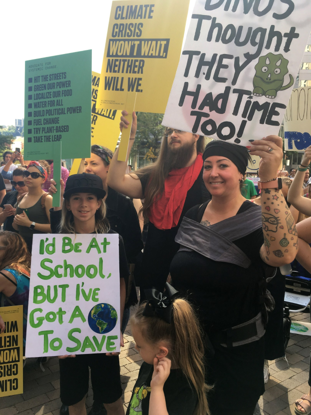 Eine Gruppe von Menschen mit Schildern, die gegen den Klimawandel protestieren, vor einer Menge, mit Bäumen, Gebäuden und einem klaren blauen Himmel im Hintergrund.