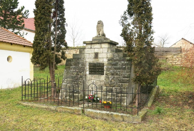 Steinernes Holocaust-Denkmal in einer grünen Wiese, umgeben von einem Metallzaun mit Blumensträußen, vor Bäumen, Häusern, einer Wand und einem klaren blauen Himmel.