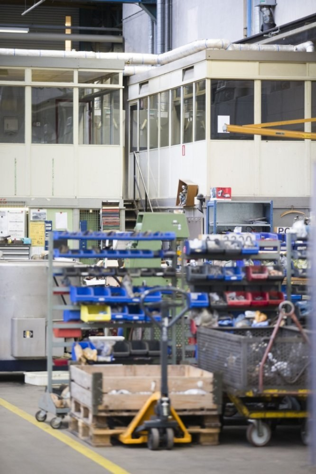 Ein großer Lagerraum voller verschiedener Geräte, einschließlich Wagen, Schränken und einer Kiste, mit einer Wand aus Glasfenstern und Rohren im Hintergrund.