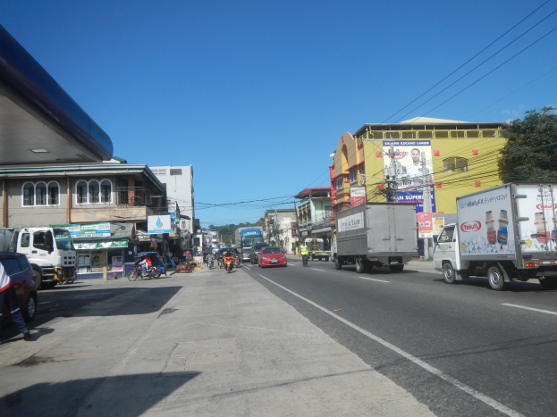 Eine belebte Stadtstraße mit zahlreichen Autos, Lastwagen, Fußgängern auf dem Gehweg, Gebäuden, Schildern, Strommasten mit Drähten, Bäumen und einem bewölkten Himmel.