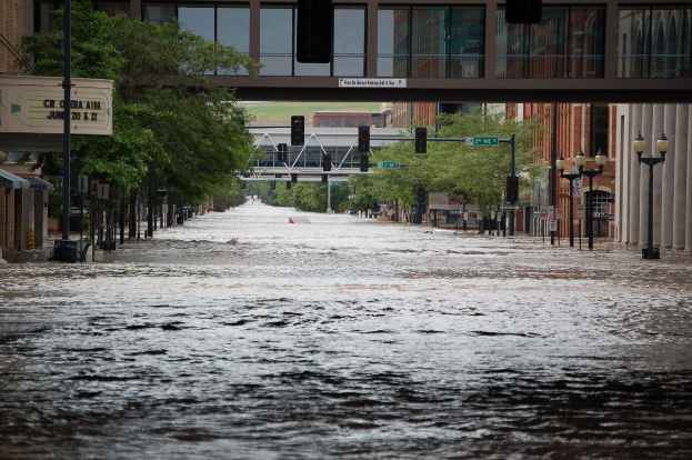 Eine überflutete städtische Straße mit Wasser, das die Straße, die Infrastruktur und die umliegenden Gebäude bedeckt, einschließlich einer Brücke im Hintergrund.