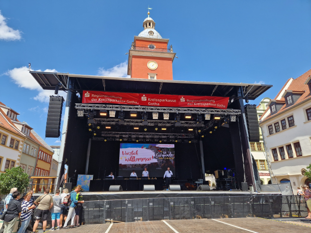 Gruppe von Menschen auf einer Bühne vor einem Uhrenturm, mit Gebäuden, Bäumen und einem bewölkten Himmel im Hintergrund, beim Kreissparkasse Gotha Musikfestival.