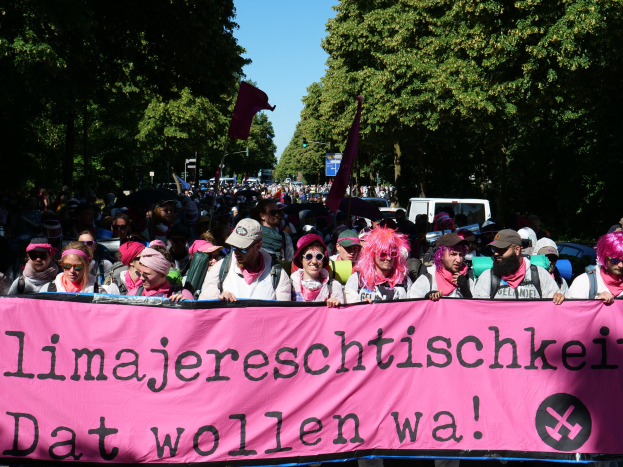 Eine Gruppe von Menschen marschiert mit einer pinken Fahne mit der Aufschrift 'Frauenmarsch in Berlin' die Straße entlang, einige tragen Mötzen und Schutzbrillen, andere halten Fahnen, mit Bäumen und Fahrzeugen im Hintergrund.
