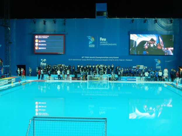 Gruppe von Menschen, die um ein Hallenbad mit Netz am Boden stehen, mit einem Wandbildschirm im Hintergrund, der "FINA World Championships 2015 Kazan" anzeigt, sowie Träger und Beleuchtung.