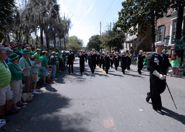 Eine Gruppe von Menschen marschiert auf einer Straße in einem St. Patrick's Day-Umzug, einige spielen Musikinstrumente, während andere von der linken Seite zuschauen; ein Gebäude, Bäume und Strommasten sind im Hintergrund zu sehen.