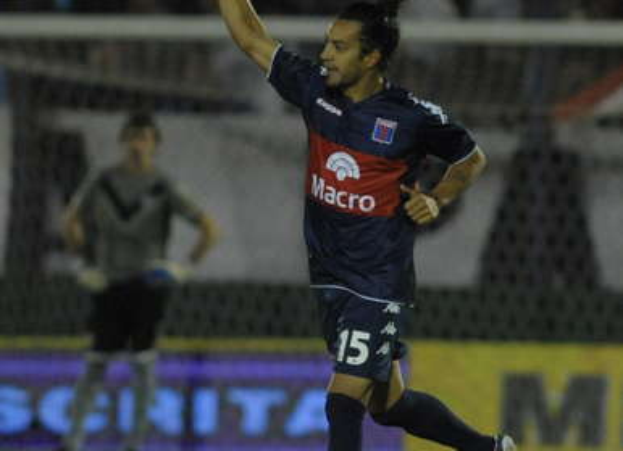 Ein Fußballspieler feiert auf dem Boden nach einem Tor, mit einer jubelnden Menge und einer Person im Hintergrund und einer Tafel mit Text dahinter, alles mit einem unscharfen, energetischen Hintergrund.