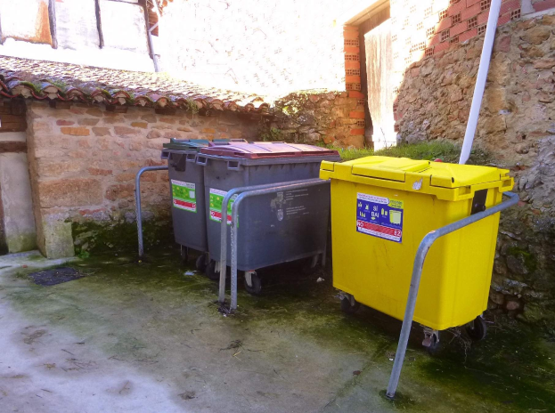 Zwei gelbe und graue Recyclingcontainer mit Rädern nebeneinander vor einem Gebäude mit einer Wand und einem Fenster im Hintergrund.