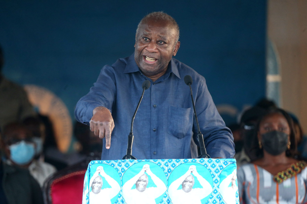 Ein Mann in einem blauen Hemd spricht in zwei Mikrofone an einem Rednerpult, mit einer Menge und mehreren sitzenden Personen, einige tragen Masken, im Hintergrund.