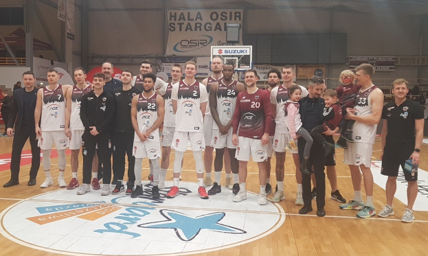 Mannergruppe auf einem Basketballfeld mit Kindern, sitzenden Zuschauern, Bannern mit "Hala Osir Starga", einem Basketballkorb, Metallstangen, Deckenlampen und einer Wand im Hintergrund.