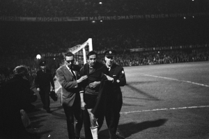 Schwarzes Bild von Fußballspielern, die auf einem Feld mit Torpfosten und beleuchteter Stadionkulisse feiern.
