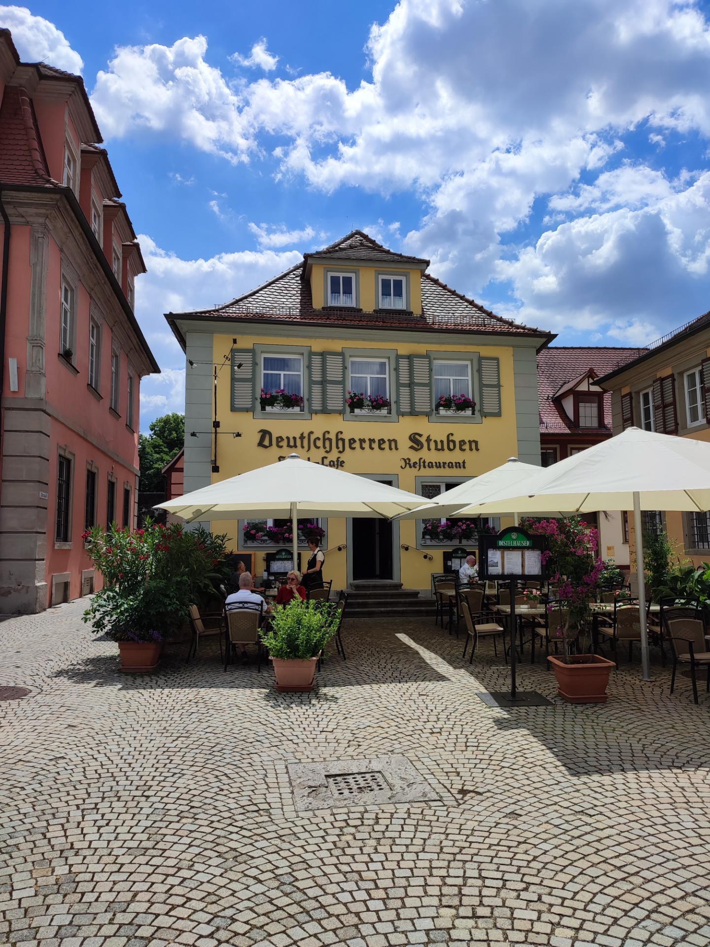 Außenbereich mit Tischen und Sonnenschirmen vor einem gelben Gebäude mit der Aufschrift "Deutschen Stuben Restaurant" in Baden-Württemberg, Deutschland, mit einigen Menschen, die sitzen und stehen, unter einem blauen und weißen Himmel.
