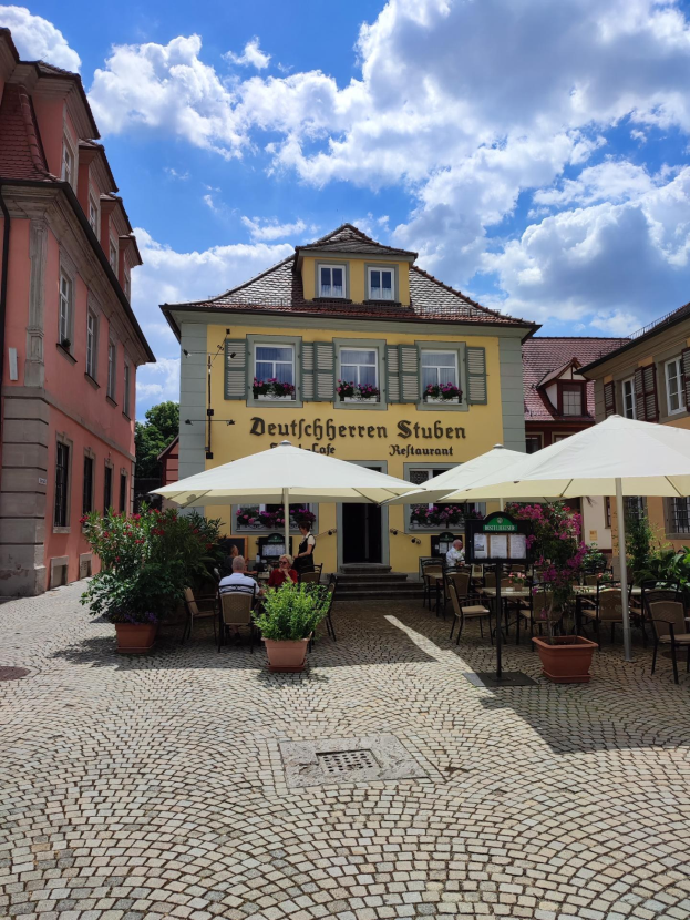 Außenbereich mit Tischen und Sonnenschirmen vor einem gelben Gebäude mit der Aufschrift "Deutschen Stuben Restaurant" in Baden-Württemberg, Deutschland, mit einigen Menschen, die sitzen und stehen, unter einem blauen und weißen Himmel.