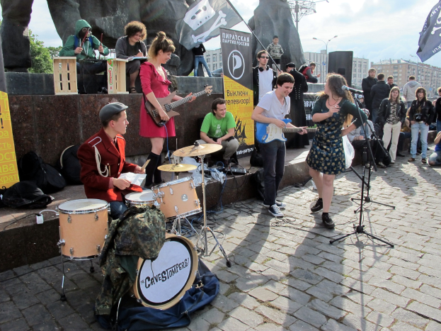 Eine Gruppe von Menschen spielt Musikinstrumente auf einem Bürgersteig mit verstreuter Ausrüstung, Bannern, einer Statue, Bäumen, Gebäuden und einem bewölkten Himmel im Hintergrund.