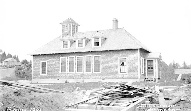 Schwarz-weiß-Foto eines zweistöckigen Gebäudes mit der Beschriftung "Regierungs-Schule, Thwaites, 1905" mit Fenstern, Türen und umgeben von Bäumen unter einem klaren Himmel, mit Holzplanken unten.