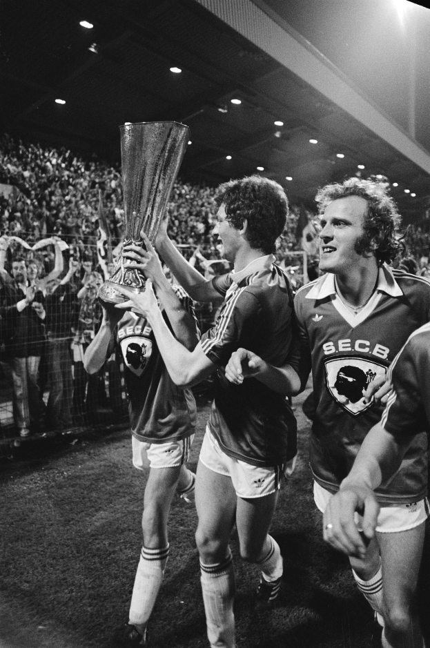 Schwarzes Bild von Fußballspielern, die mit einem Pokal auf einem Feld stehen, umgeben von einer Menge und Stadionbeleuchtung im Hintergrund.