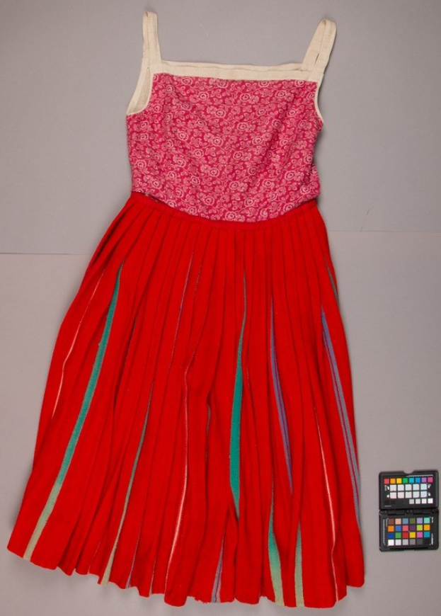 Ein rotes Kleid mit einem pinken Oberteil und grünen Streifen, das eine plissierte Röcke und einen Ausschnitt hat, liegt auf dem Boden neben einem leuchtenden Farbpalette.