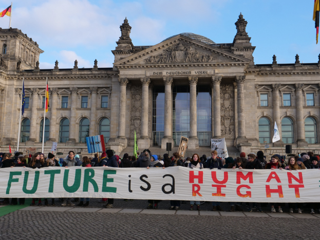 Eine Gruppe von Menschen hält ein Transparent mit der Aufschrift 'Zukunft ist ein Menschenrecht' vor dem Reichstagsgebäude in Berlin, Deutschland, mit sichtbaren Fenstern, Säulen und Bögen des Gebäudes und umgeben von Fahnenmasten gegen einen bewölkten Himmel.