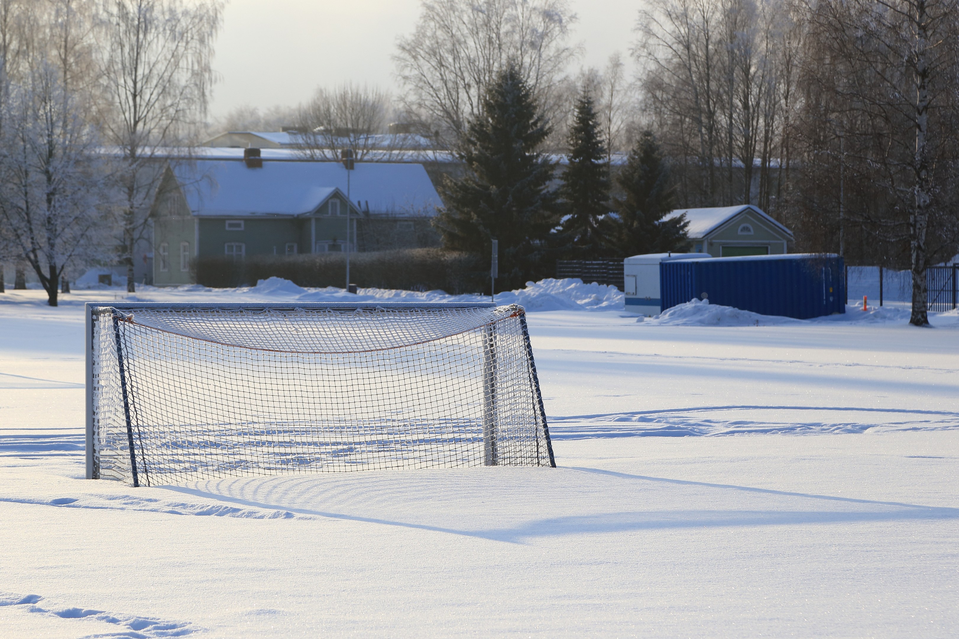 Fußballtor auf einem verschneiten Feld mit einem Netz, Pfosten, einem Zaun, einem Container, Bäumen, Häusern und einem bewölkten Himmel.