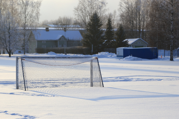 Fußballtor auf einem verschneiten Feld mit einem Netz, Pfosten, einem Zaun, einem Container, Bäumen, Häusern und einem bewölkten Himmel.
