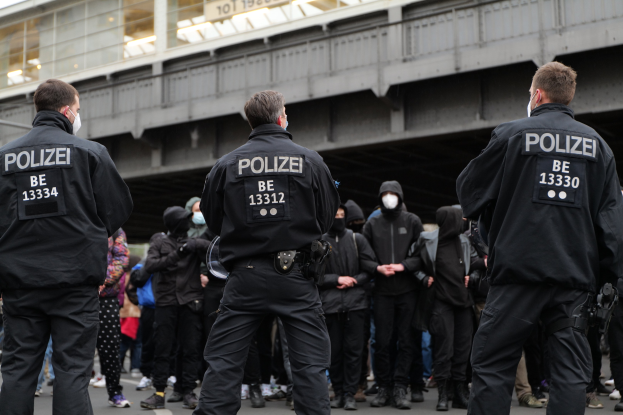 Eine Gruppe uniformierter Polizisten steht vor einer Menge von Menschen in schwarzen Uniformen und Masken, im Hintergrund eine Brücke und ein Gebäude.