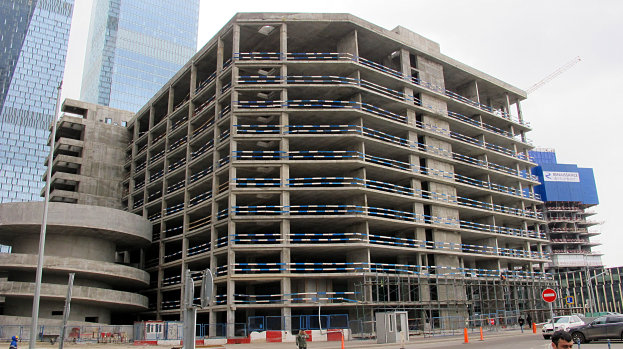 Großes Bauprojekt in einer städtischen Umgebung mit umliegenden Wolkenkratzern, Fahrzeugen, Fußgängern, Verkehrsinfrastruktur und Kränen vor einem klaren Himmel.