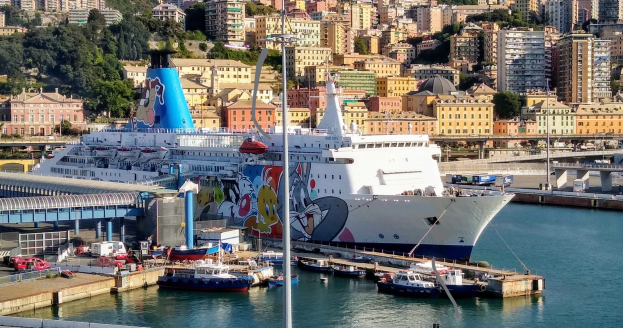 Großes Kreuzfahrtschiff in der Nähe einer Stadt mit einer Brücke links, Fahrzeugen auf der Straße und Gebäuden und Bäumen im Hintergrund.