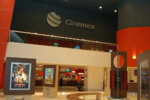 Eine Gruppe von Menschen in einem Cinemex-Kino in einem Einkaufszentrum, mit einer Person auf einem Stuhl, einem Tisch, Fotos auf der Wand, einem Schild, Deckenleuchten und Geräten auf den Tischen.