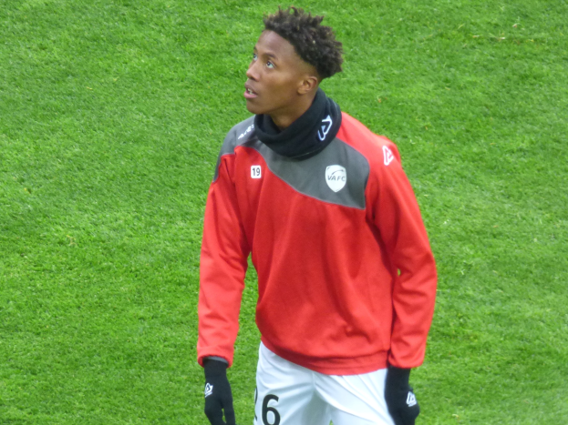 Ein junger Mann in einer roten Jacke und weißen Shorts steht auf einem Fußballfeld, trägt Baseball-Handschuhe und sieht entschlossen nach rechts.