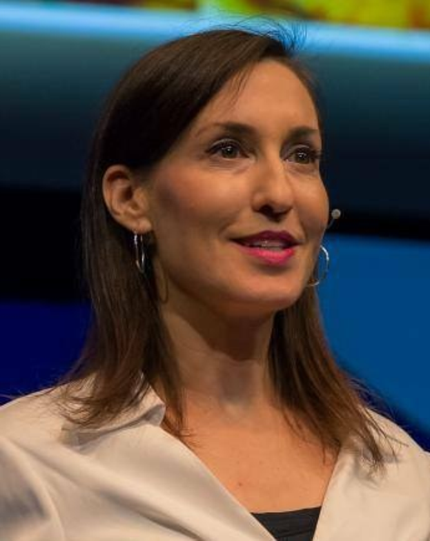 Frau in weißer Jacke und schwarzem Oberteil, die für ein Interview sitzt, mit Ohrringen und Mikrofon, vor einem blauen und gelben Hintergrund.