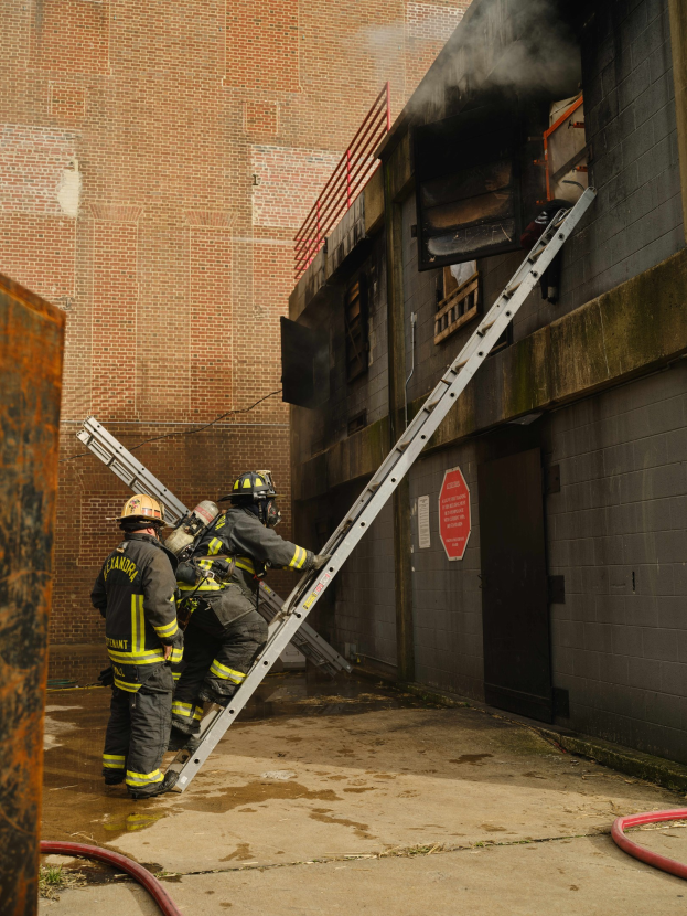 Feuerwehrleute in Helmen klettern eine Leiter zu einem rauchgefüllten Gebäude hoch, mit einem Metallobjekt links und Rohren am Boden.