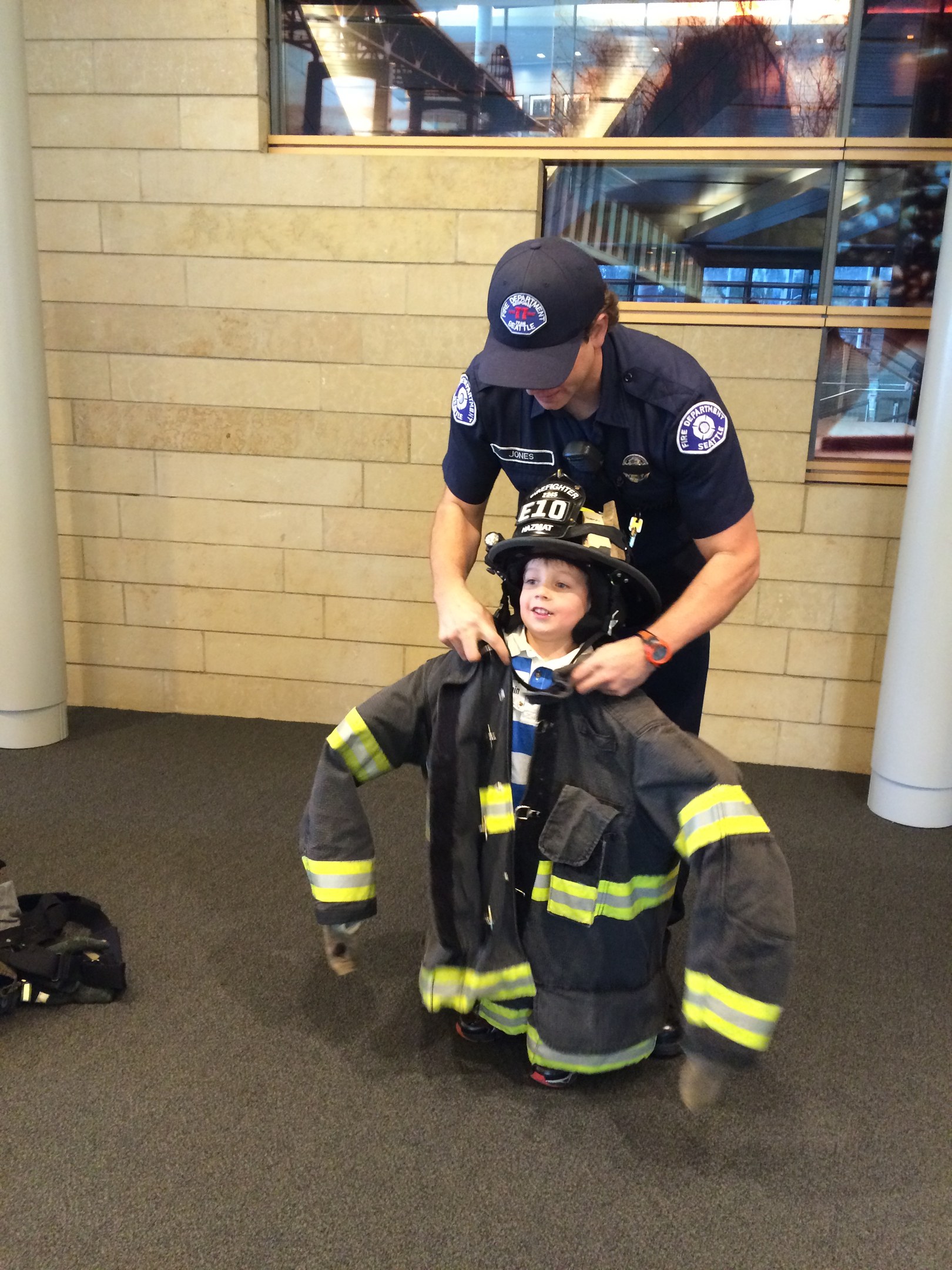 Ein junger Junge in Feuerwehruniform wird von einem Polizeibeamten unterstützt, mit Taschen auf dem Boden neben ihnen und einer Brückenreflexion im Hintergrund durch eine Glasscheibe sichtbar.