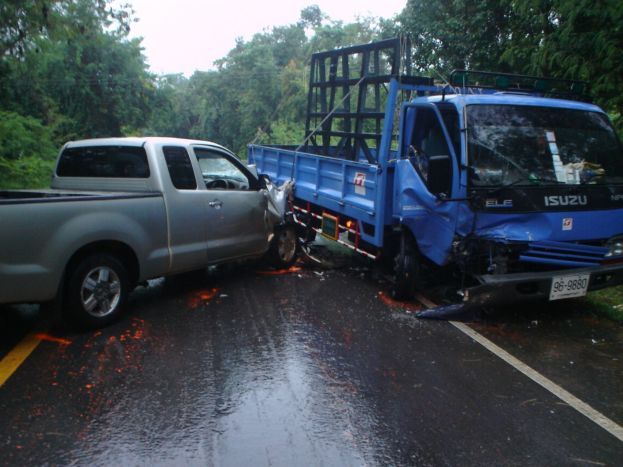 Ein schwer beschädigtter Lkw mit eingedellter Front und verbeulter Karosserie liegt am Straßenrand neben Bäumen unter einem klaren blauen Himmel.