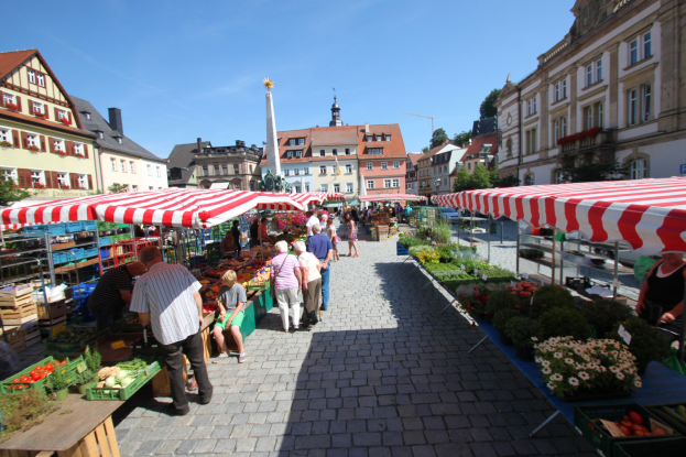 Ein belebter Markt im historischen Stadtkern von Heidelberg mit Menschen, die spazieren gehen, auf Bänken sitzen und in der Nähe von Zelten stehen, mit Gemüsekörben auf Tischen, umgeben von Gebäuden, Bäumen und einem klaren blauen Himmel.