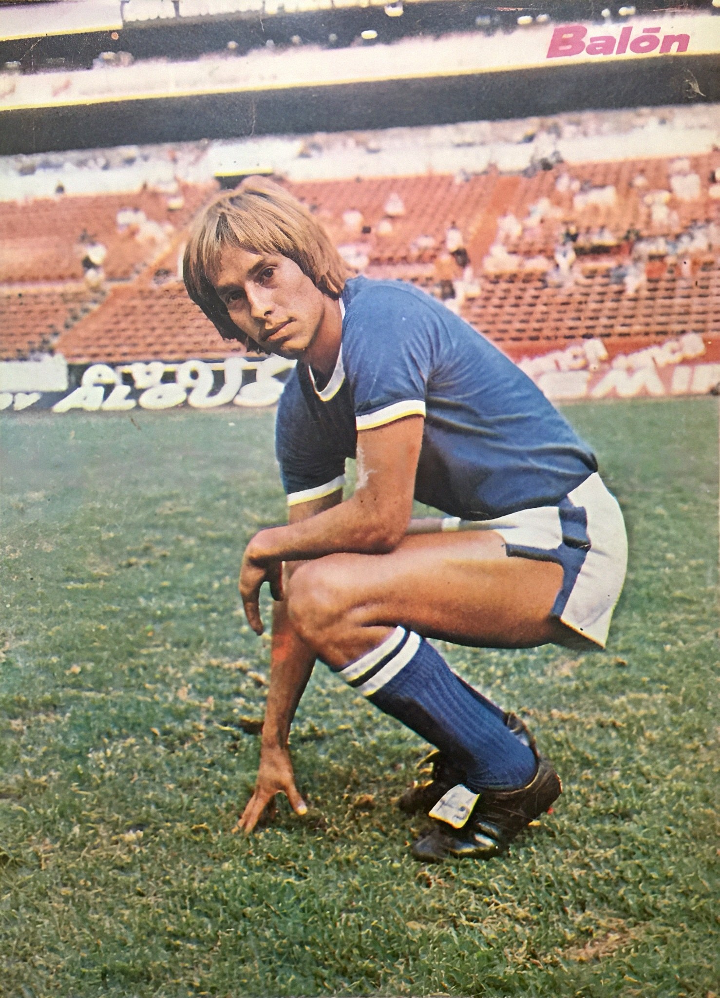 Ein Mann kniet auf einem Fußballfeld umgeben von Menschen mit Schildern und trägt Sportschuhe.